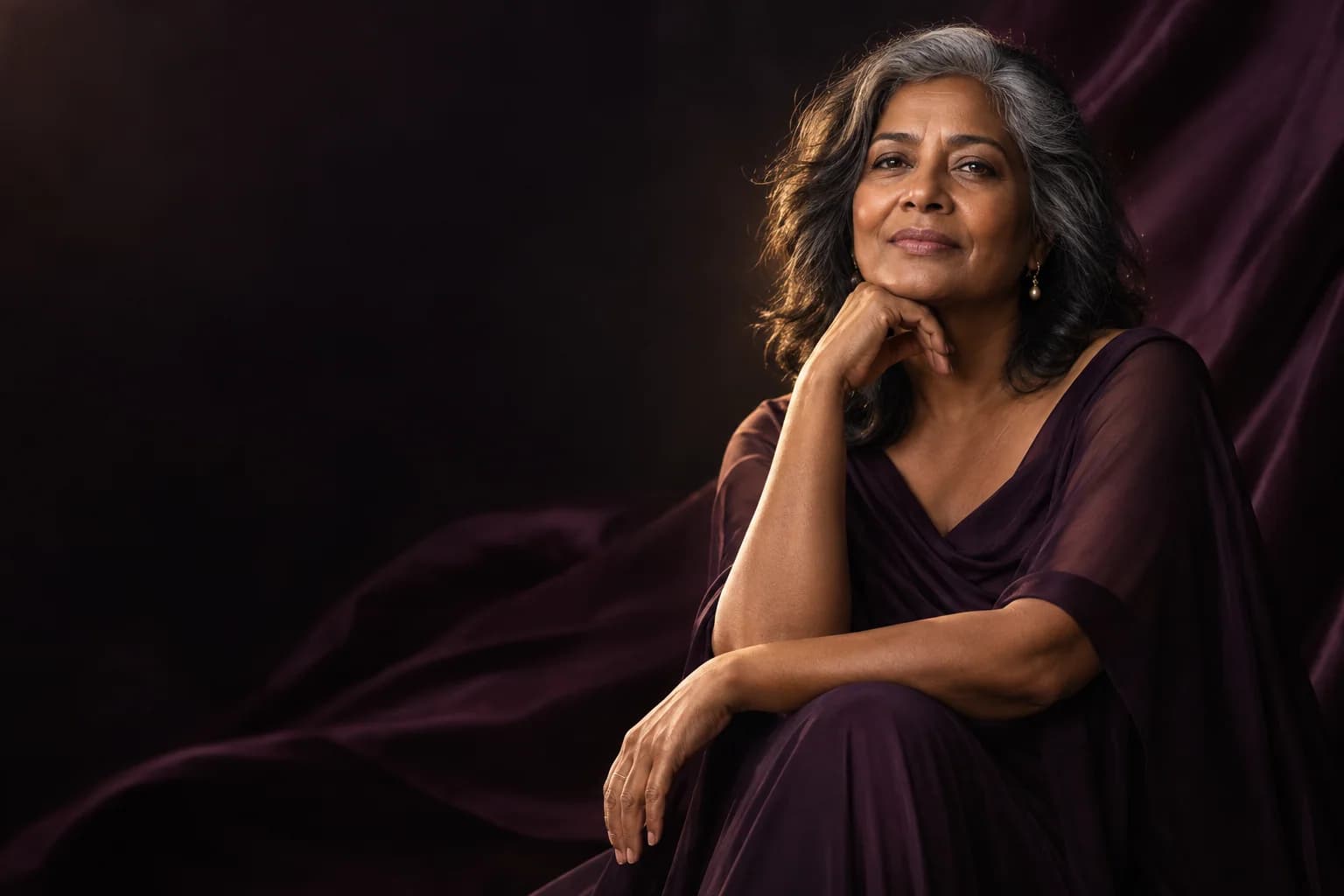 A confident mature woman seated against deep-plum drapery.